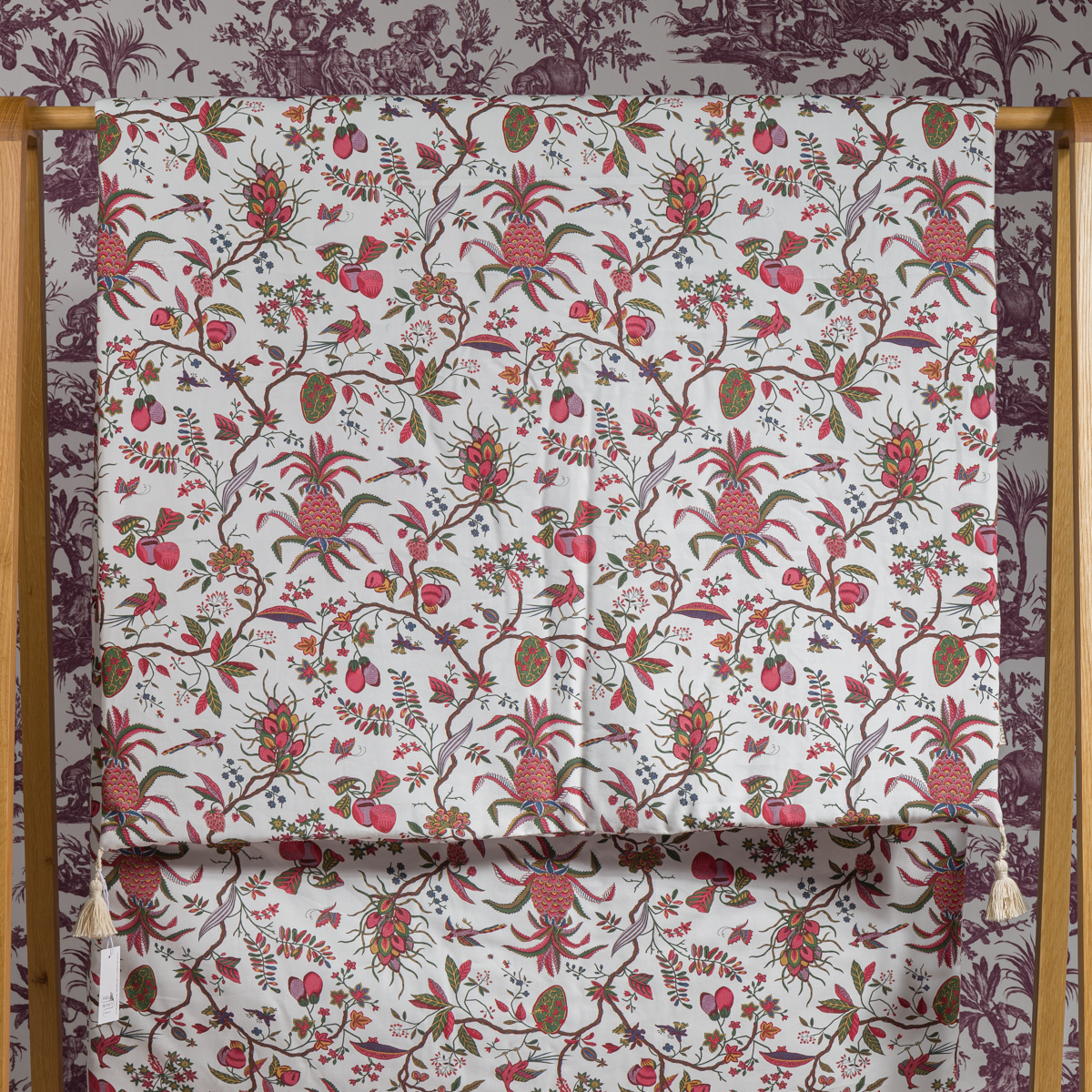 Le MATELAS DE REPOS "ANANAS", un tissu aux motifs botaniques rouges, roses et verts d'ananas, de grenades et de fleurs, est drapé sur un cadre en bois avec un papier peint toile coordonné en arrière-plan. la marque Toile de Jouy