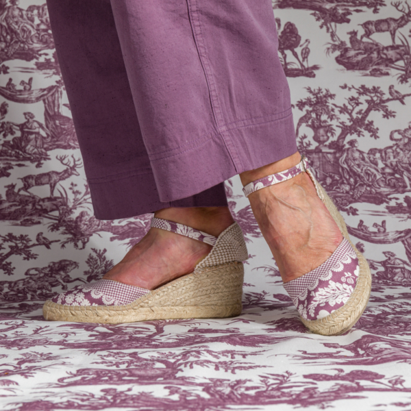 Une personne portant un pantalon lavande et des "ESPADRILLES à BRIDE et TALON 'FLEURS STYLISéES' AMARANTE" avec semelles en jute se tient sur un tissu de toile violet et blanc assorti. la marque Toile de Jouy