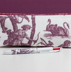 Le MATELAS DE REPOS "FLEURS STYLISÉES" MANDARINE présente un stylo blanc à motifs violets reposant devant une pochette en tissu décorée de toile violette, représentant un personnage avec un nœud, deux cygnes, le tout sur un fond blanc et violet. la marque Toile de Jouy