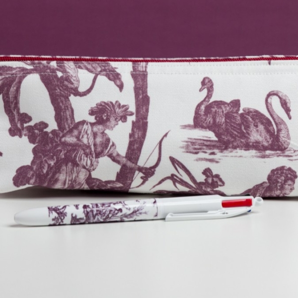 Le MATELAS DE REPOS "FLEURS STYLISÉES" MANDARINE présente un stylo blanc à motifs violets reposant devant une pochette en tissu décorée de toile violette, représentant un personnage avec un nœud, deux cygnes, le tout sur un fond blanc et violet. la marque Toile de Jouy