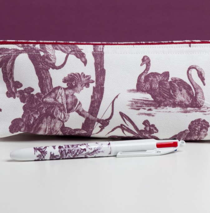Le MATELAS DE REPOS "FLEURS STYLISÉES" MANDARINE présente un stylo blanc à motifs violets reposant devant une pochette en tissu décorée de toile violette, représentant un personnage avec un nœud, deux cygnes, le tout sur un fond blanc et violet. la marque Toile de Jouy