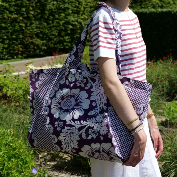 Vêtue d'un t-shirt rayé rouge et blanc et d'un pantalon blanc, une personne se tient debout dans un jardin, un grand sac fourre-tout à motifs floraux violets, le « XCOMM_SAC SHOPPING FLEURS AMARANTE », en bandoulière. la marque Toile de Jouy