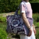 Vêtue d'un t-shirt rayé rouge et blanc et d'un pantalon blanc, une personne se tient debout dans un jardin, un grand sac fourre-tout à motifs floraux violets, le « XCOMM_SAC SHOPPING FLEURS AMARANTE », en bandoulière. la marque Toile de Jouy
