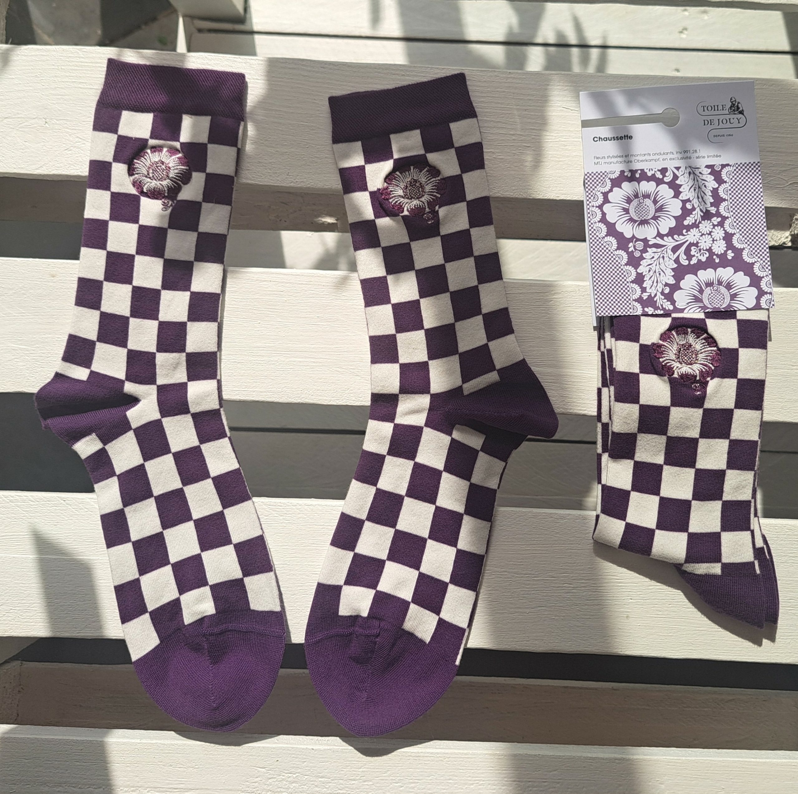 Trois paires de chaussettes XCOMM_CHAUSETTES FLEURS STYLISÉES AMARANTE — à carreaux violets et blancs ornées d’un motif floral — sont disposées sur une surface en bois clair. Une paire se trouve dans son emballage, à droite, et la lumière du soleil projette de douces ombres sur la scène. la marque Toile de Jouy