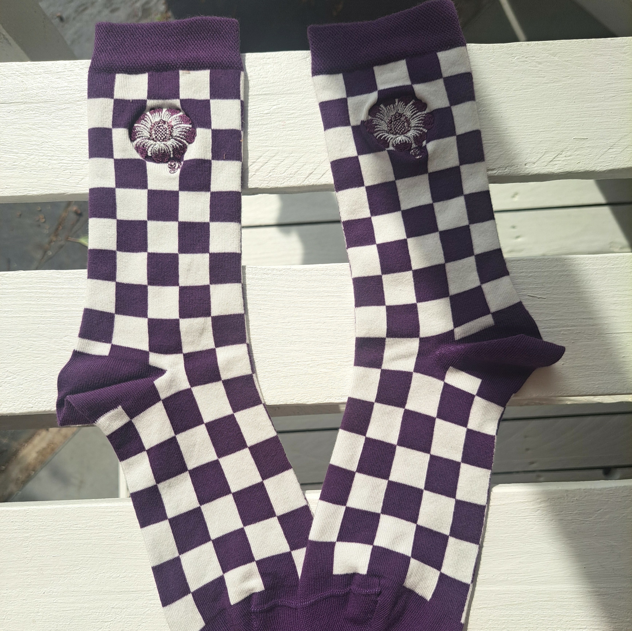 Une paire de chaussettes XCOMM_CHAUSETTES FLEURS STYLISÉES AMARANTE, ornées d'un motif à carreaux violets et blancs et d'une fleur brodée blanche près du bord supérieur, posées à plat sur une surface en bois blanche, à la lumière du soleil. la marque Toile de Jouy