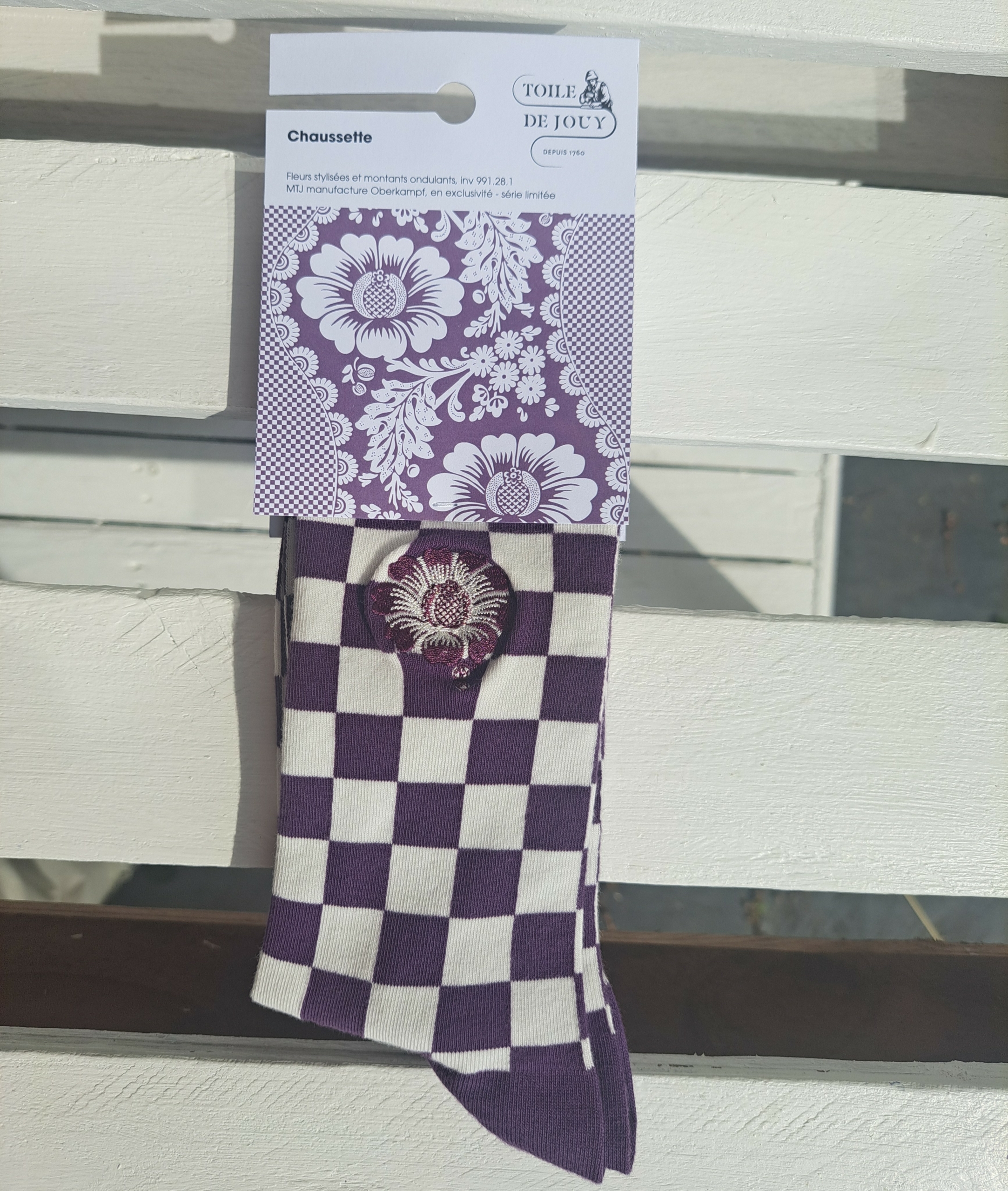 Une paire de chaussettes XCOMM_CHAUSETTES FLEURS STYLISÉES AMARANTE, ornées d'un motif à carreaux violets et blancs avec un emblème floral et munies d'une étiquette à imprimé floral « Toile de Jouy », est suspendue sur une surface à lattes blanches, baignée par la lumière du soleil. la marque Toile de Jouy