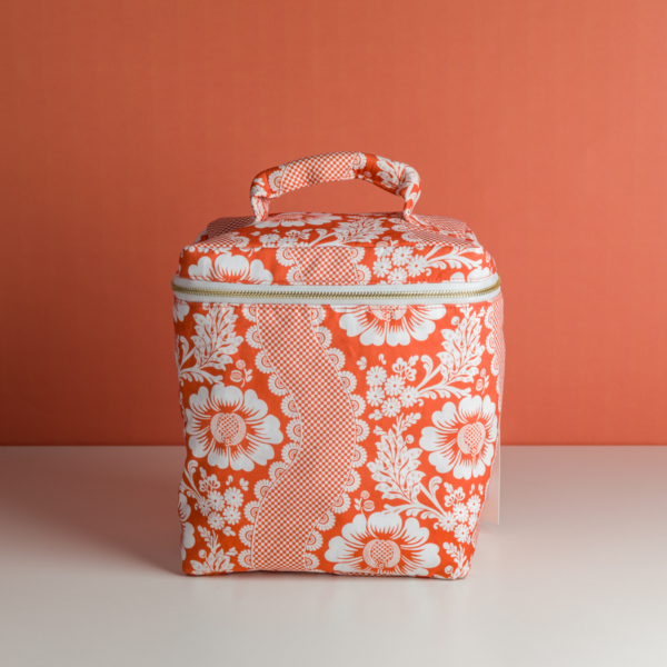 Le modèle XCOMM_VANITY FLEURS AMARANTE est un sac carré muni d'une fermeture à glissière et d'une poignée, orné d'un motif floral orange et blanc qui se détache sur un fond orange. la marque Toile de Jouy