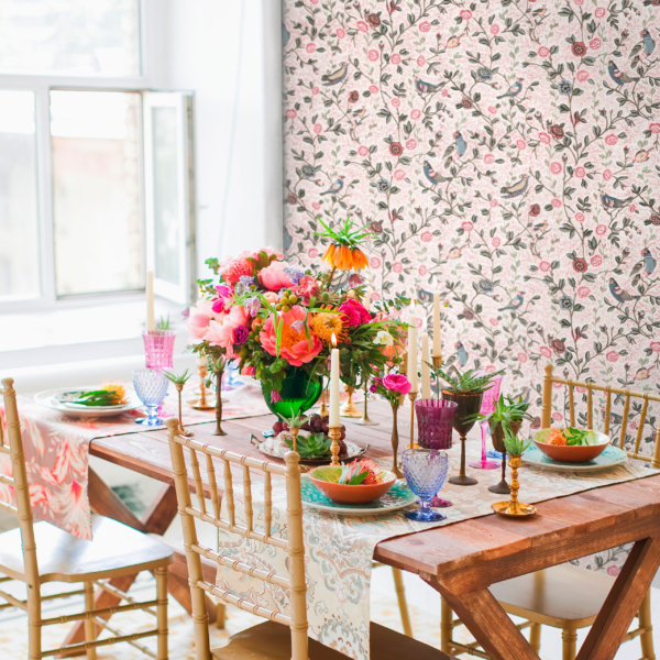 JardinMoghol-Couleur-Ambiance copie Une table à manger en bois dressée pour quatre personnes, avec des assiettes colorées, des verres, des compositions florales et des bougies. En arrière-plan, on aperçoit un papier peint à motifs floraux et une fenêtre baignée de lumière. L'ambiance est joyeuse et festive. la marque Toile de Jouy