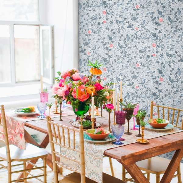 JardinMoghol-Porcelaine-Ambiance copie Une table en bois dressée pour quatre personnes, avec des assiettes à motifs floraux, des verres colorés, des chaises dorées et un bouquet éclatant en centre de table, le tout devant une fenêtre et un papier peint à motifs floraux. la marque Toile de Jouy