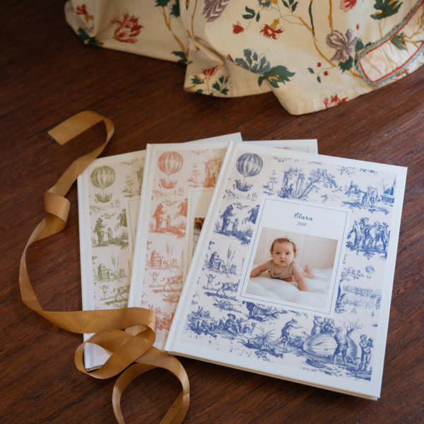 OTH_8041edited. Trois albums photo aux couvertures illustrées de toile, empilés sur un parquet, avec un ruban doré et un tissu à motifs floraux en arrière-plan. la marque Toile de Jouy
