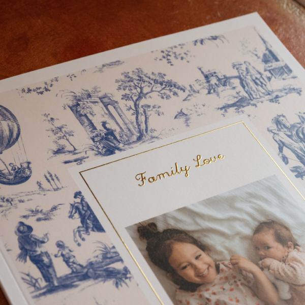 OTH_8075 Un tirage photo orné d'une bordure à motif « toile de Jouy » bleu et blanc, avec l'inscription « Family Love » en feuille d'or au-dessus d'une photo représentant une jeune fille et un bébé. la marque Toile de Jouy